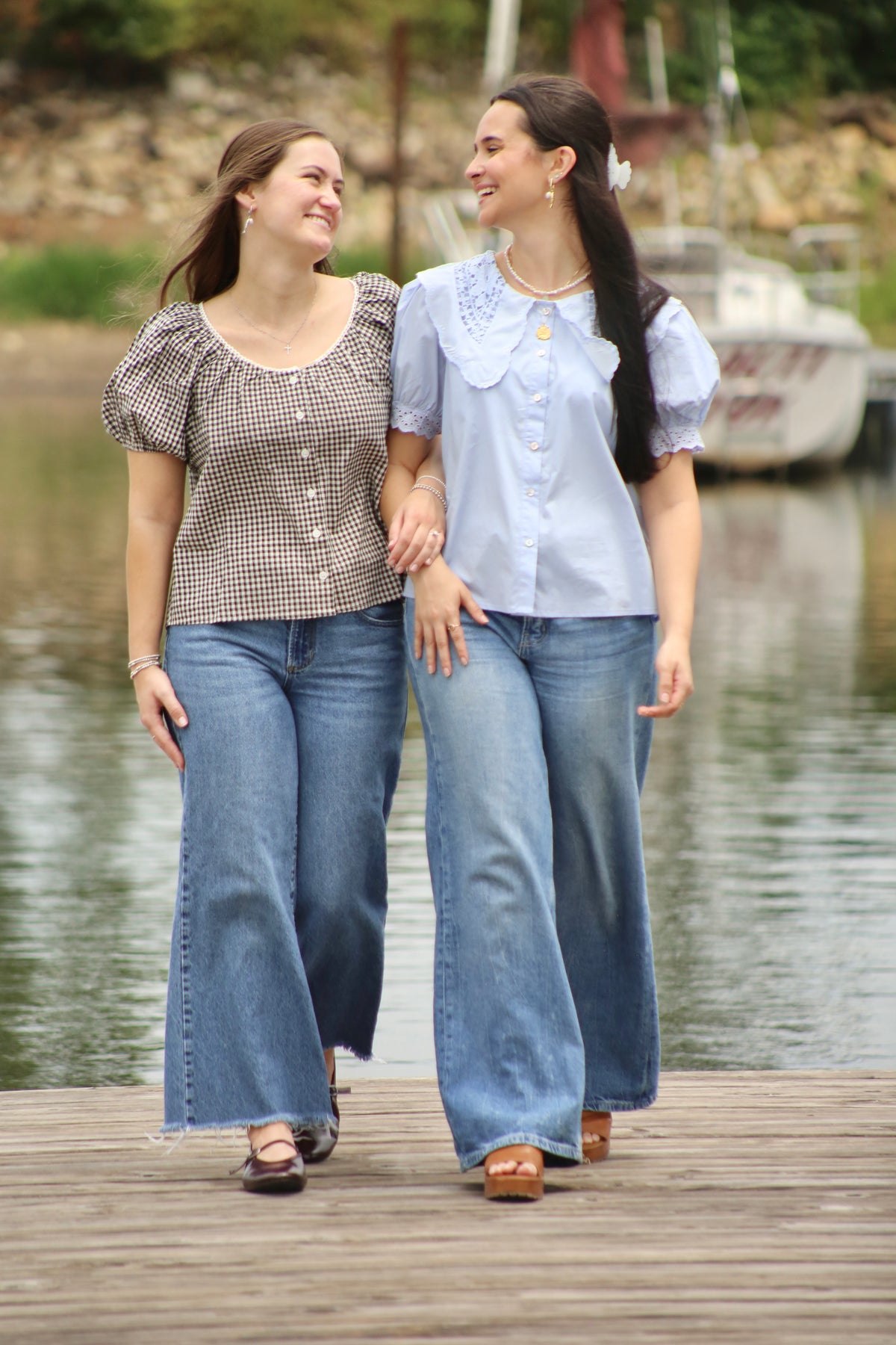 Two women walking together on a lakeside dock wearing blue jeans, one in a light blue lace-collar blouse and the other in a brown gingham puff-sleeve top.