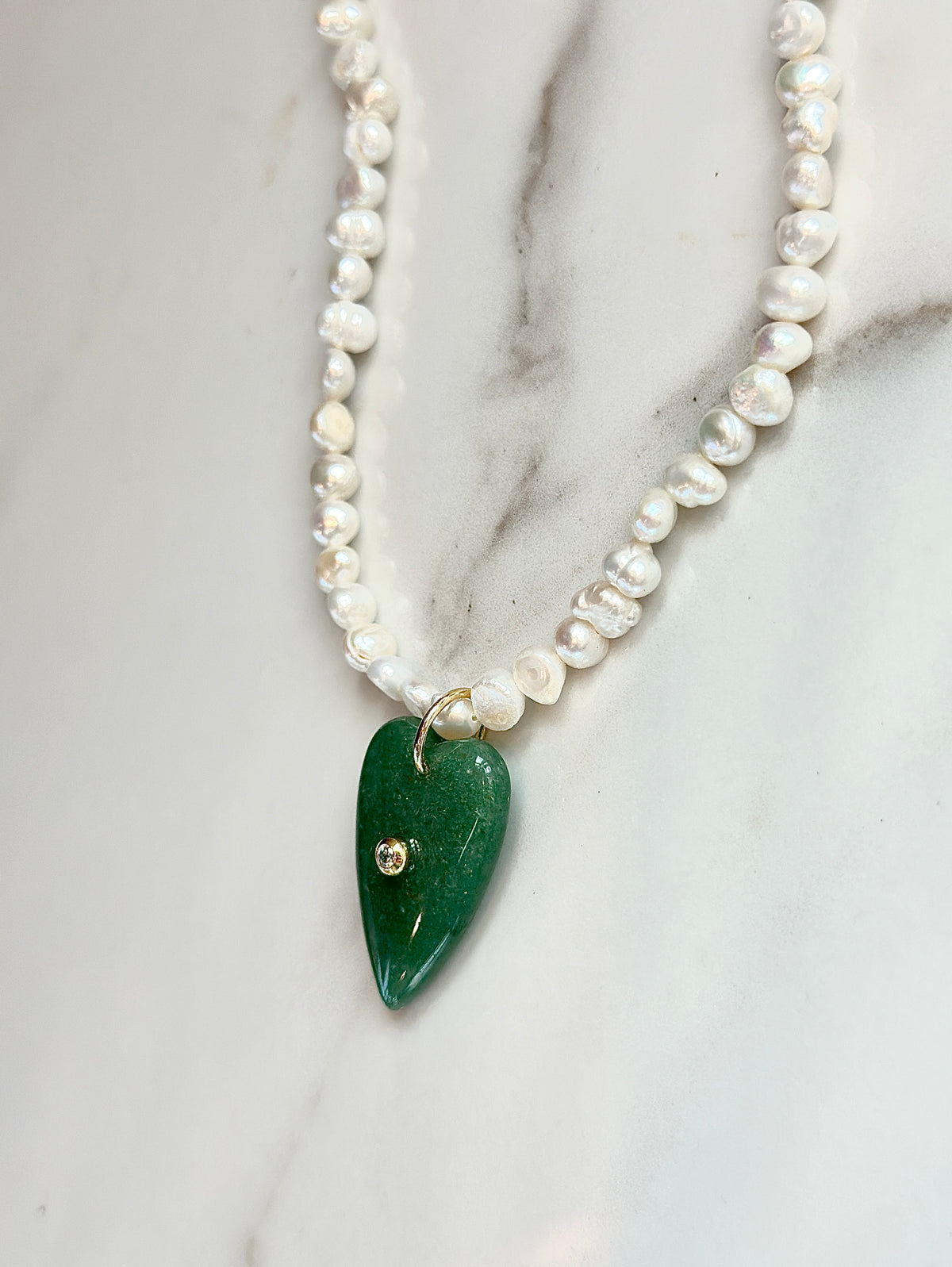 A freshwater pearl beaded necklace featuring irregular white baroque pearls and a polished green teardrop pendant with a small gold stud detail, displayed on a white marble surface.