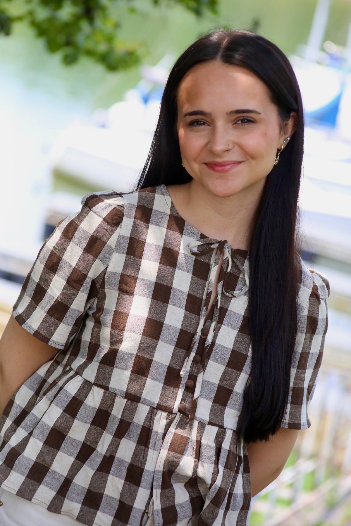 Close-up of a woman wearing a brown and cream gingham peplum blouse with puffed short sleeves, front buttons, and a tie neckline at a lakeside marina.