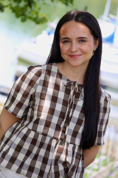 Close-up of a woman wearing a brown and cream gingham peplum blouse with puffed short sleeves, front buttons, and a tie neckline at a lakeside marina.