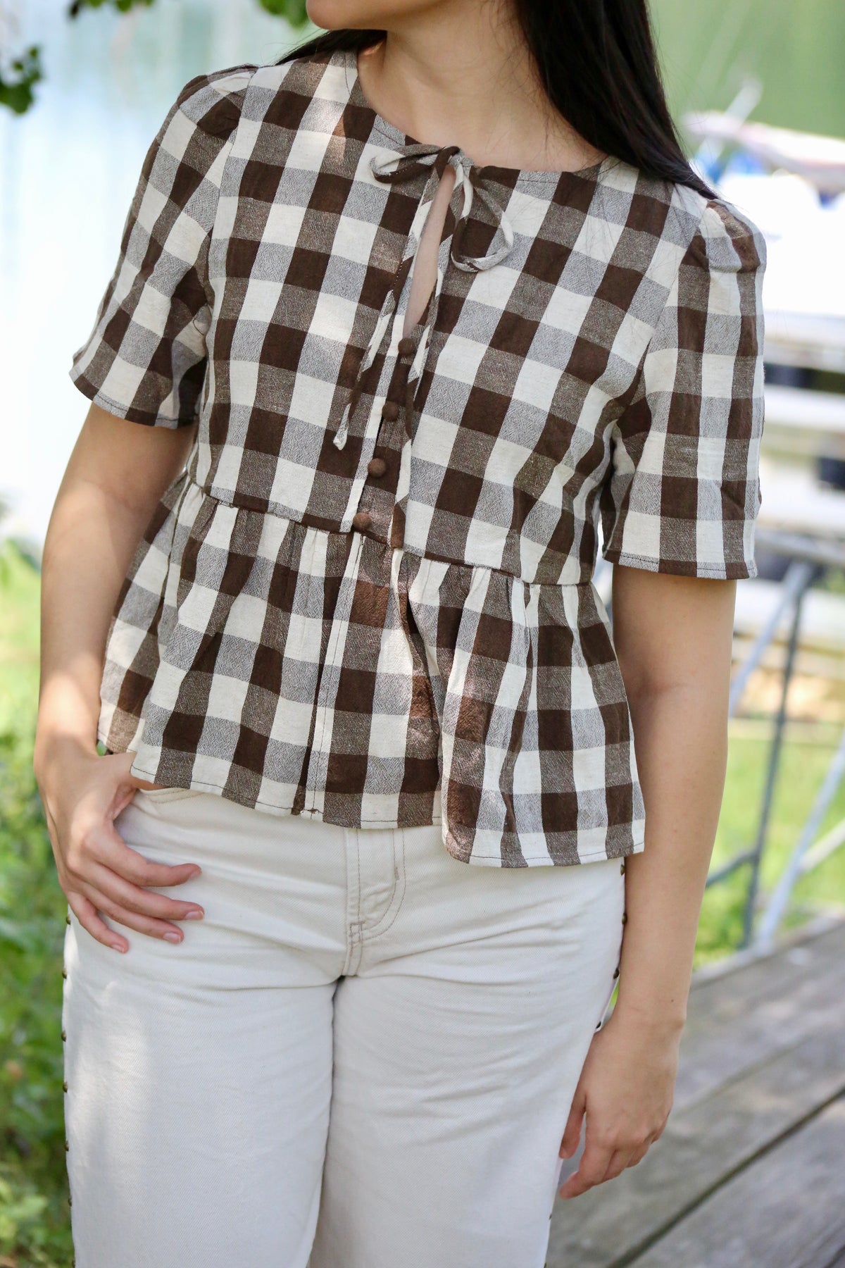 Close-up of a brown and cream gingham peplum blouse featuring short puff sleeves, front buttons, and a tie neckline, styled with cream pants.