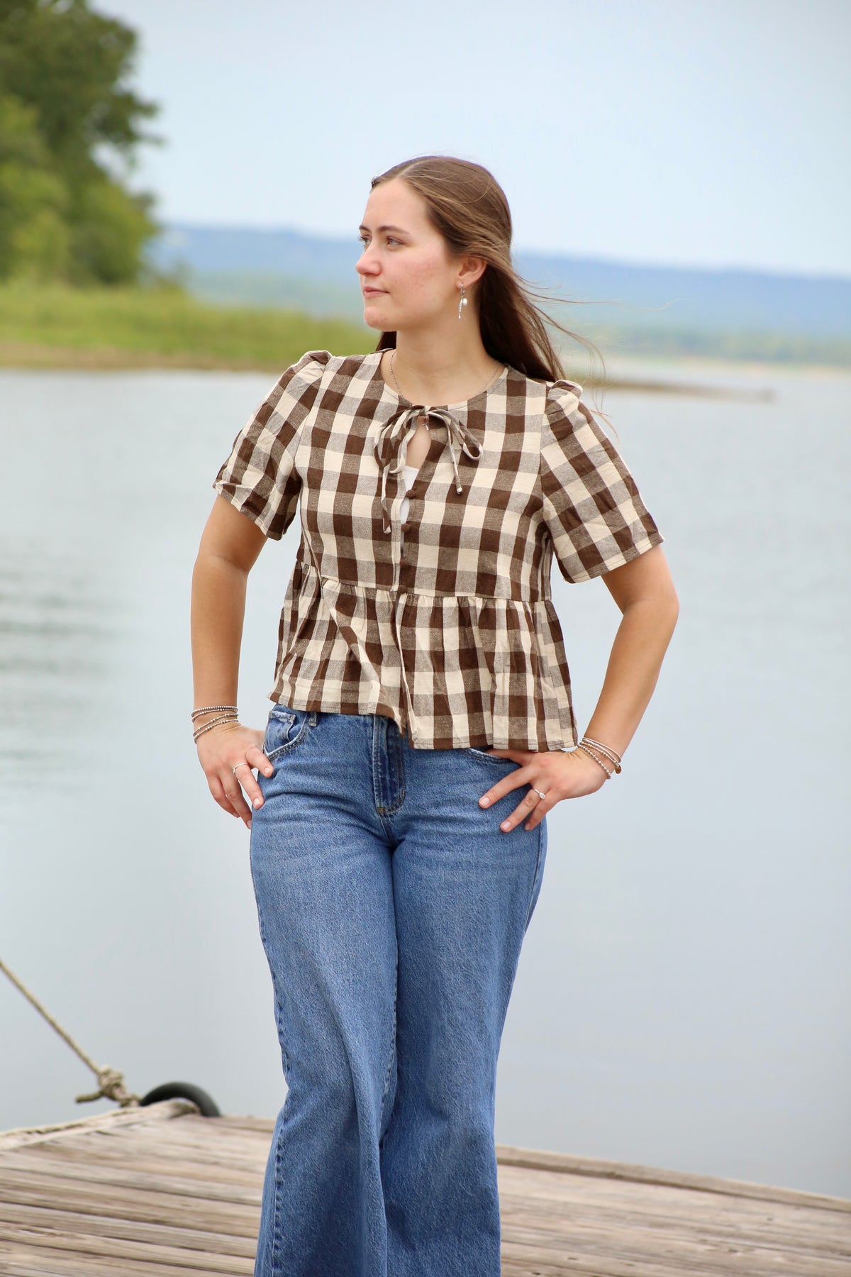 Woman wearing a brown and cream gingham peplum blouse with short sleeves and a tie-front neckline, styled with blue jeans by the lake.