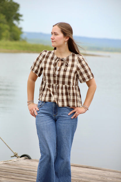 Woman wearing a brown and cream gingham peplum blouse with short sleeves and a tie-front neckline, styled with blue jeans by the lake.