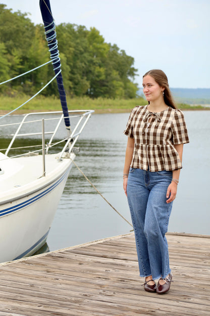Woman wearing a brown and cream gingham peplum blouse with puffed short sleeves and a tie neckline, styled with blue jeans beside a sailboat on a lakeside dock.