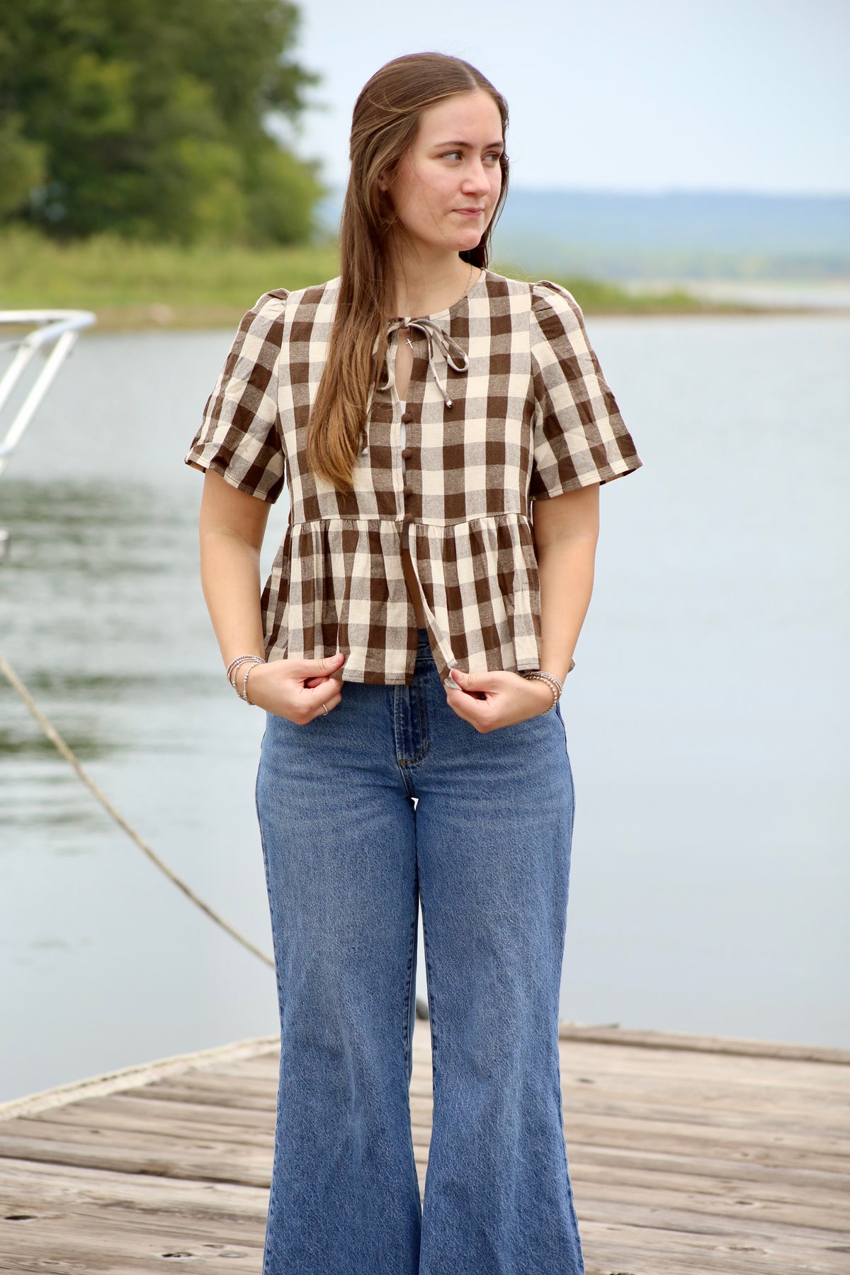 Woman wearing a brown and cream gingham peplum blouse with puffed short sleeves, front buttons, and a tie neckline, styled with blue jeans by the lake.