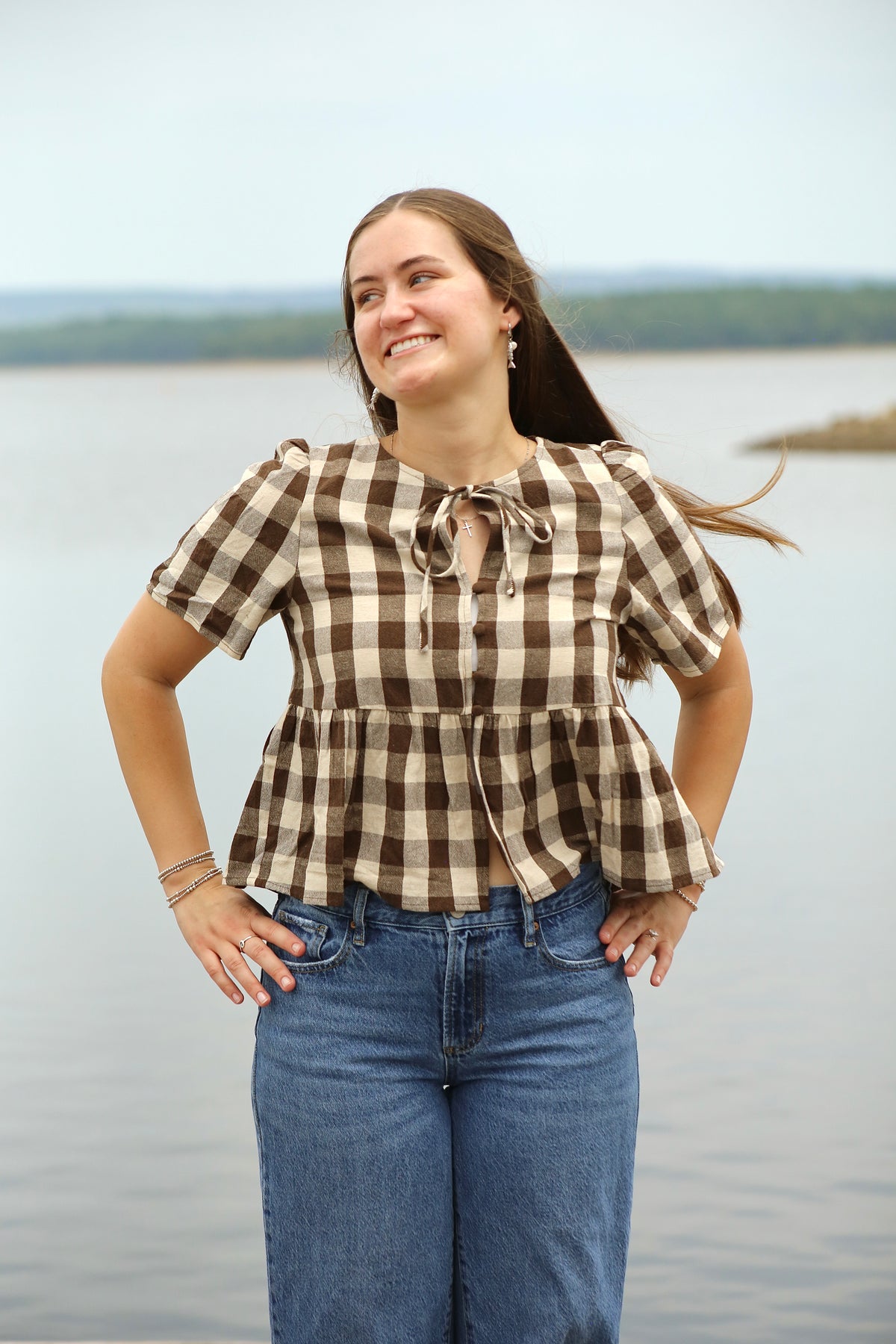 Woman wearing a brown and cream gingham peplum blouse with puffed short sleeves, front buttons, and a tie neckline, styled with blue jeans by the lake.
