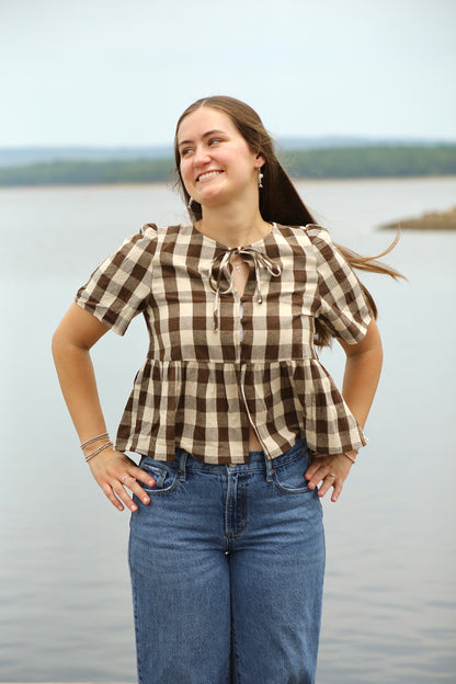 Woman wearing a brown and cream gingham peplum blouse with puffed short sleeves, front buttons, and a tie neckline, styled with blue jeans by the lake.
