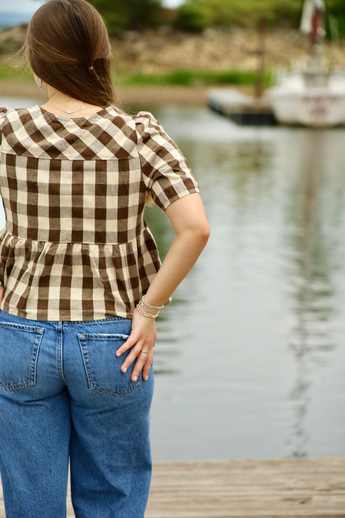 Back view of a brown and cream gingham peplum blouse with short puff sleeves, styled with blue jeans beside a lake.