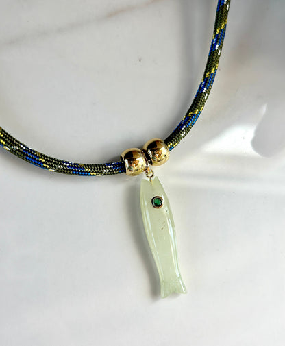 Close-up of natural green jade fish pendant with bezel-set CZ eye and gold details on patterned cord necklace.
