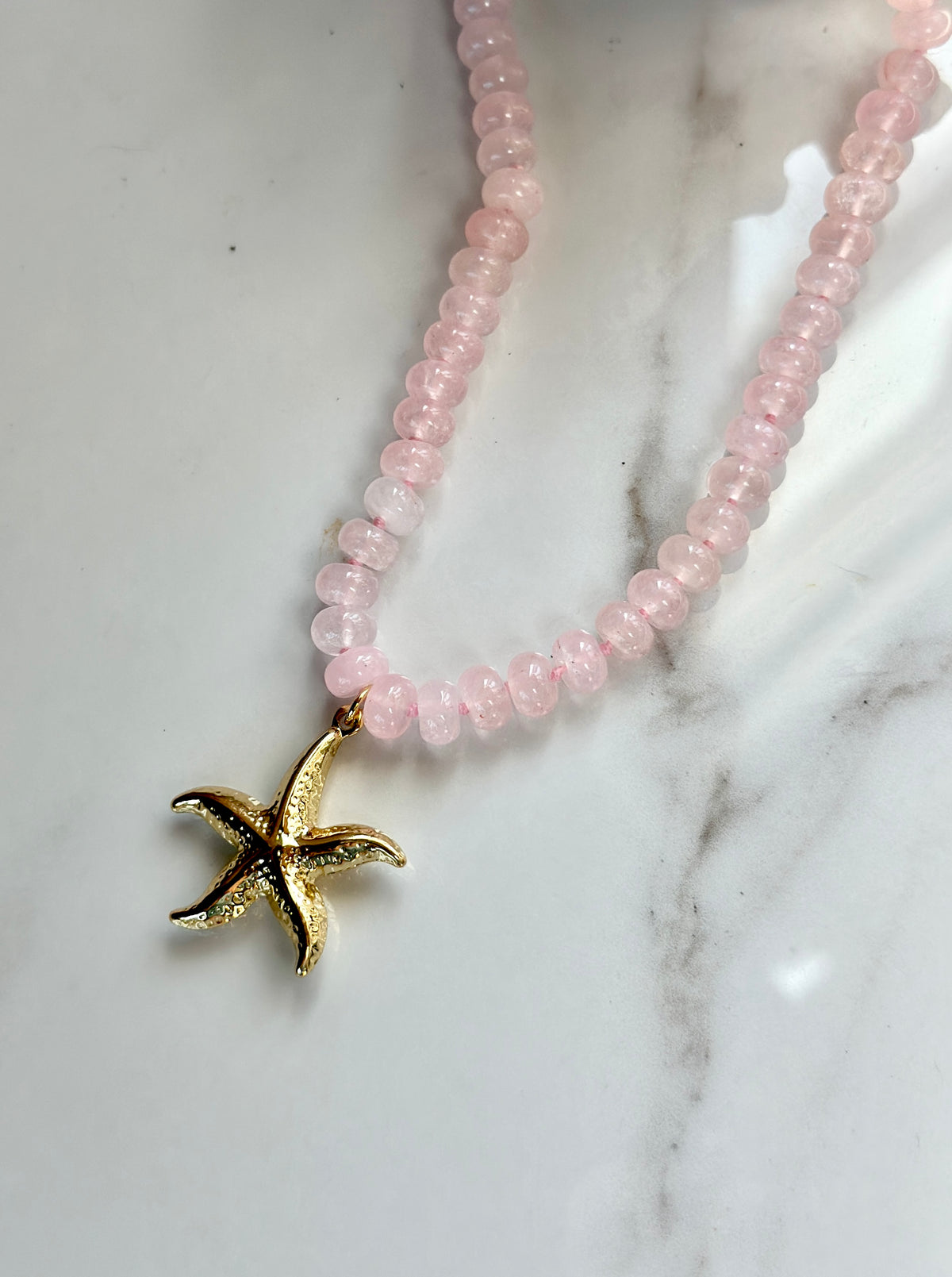 Pink gemstone starfish necklace with gold pendant displayed on white marble.
