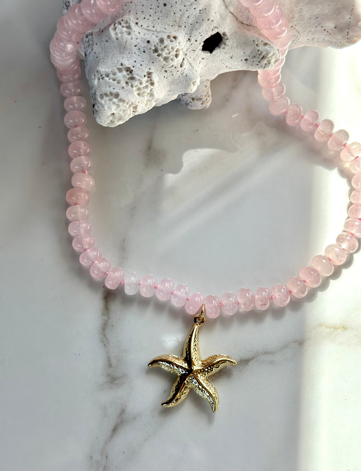 Pink natural gemstone necklace with gold starfish pendant styled over white coral.
