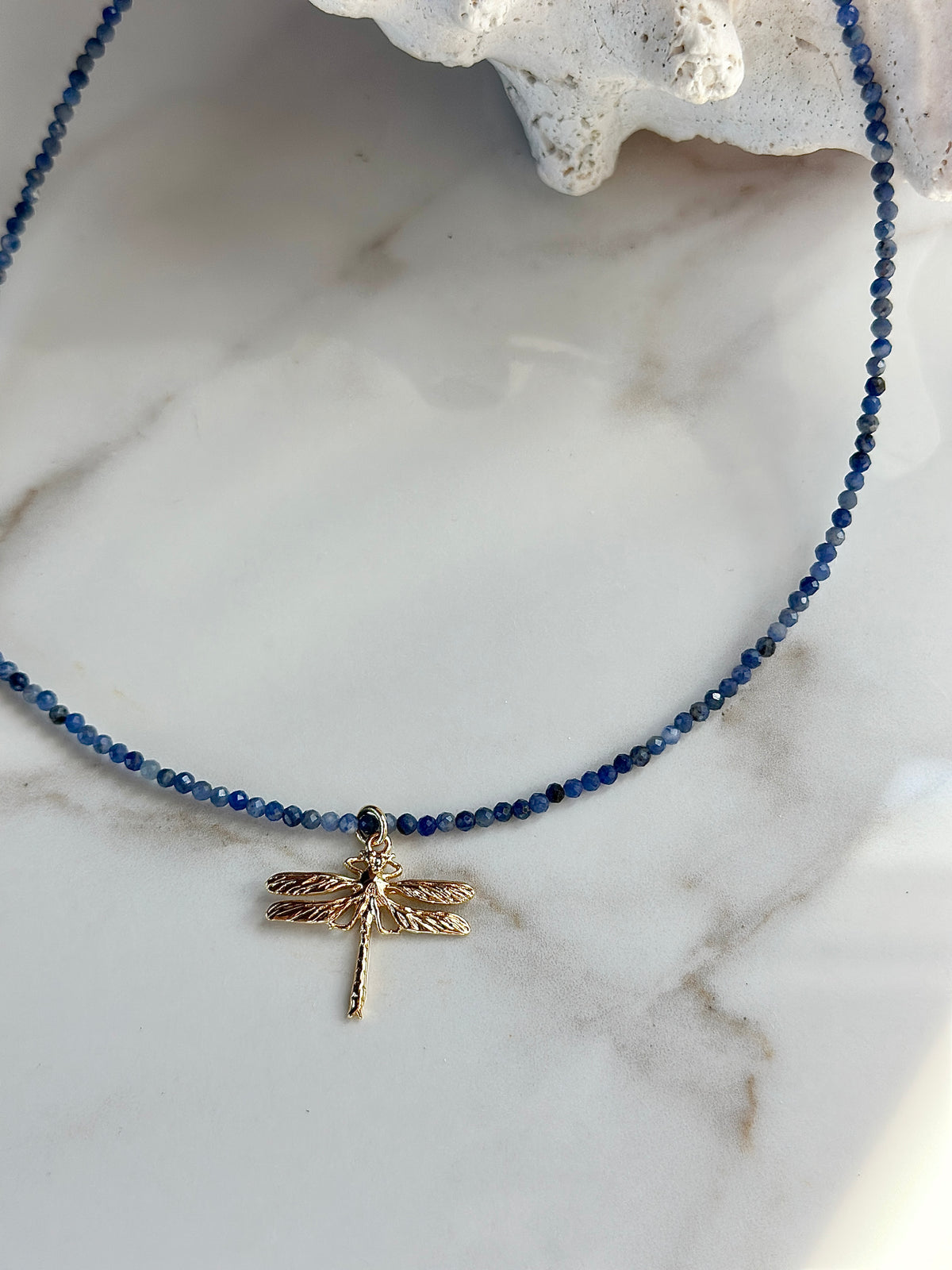 A beaded necklace with small dark blue beads and a gold dragonfly pendant, displayed on a white marble surface with a white coral fragment prop.