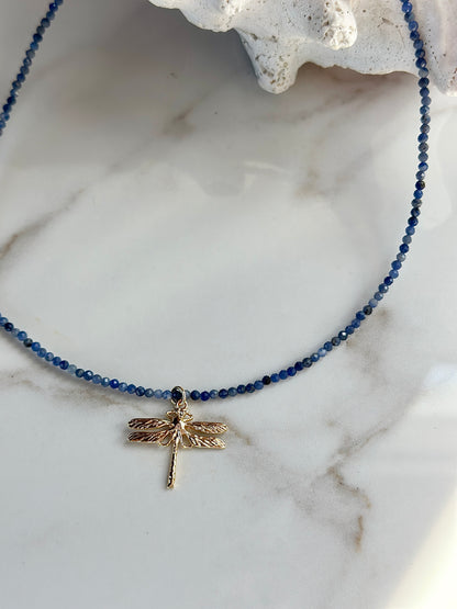A beaded necklace with small dark blue beads and a gold dragonfly pendant, displayed on a white marble surface with a white coral fragment prop.