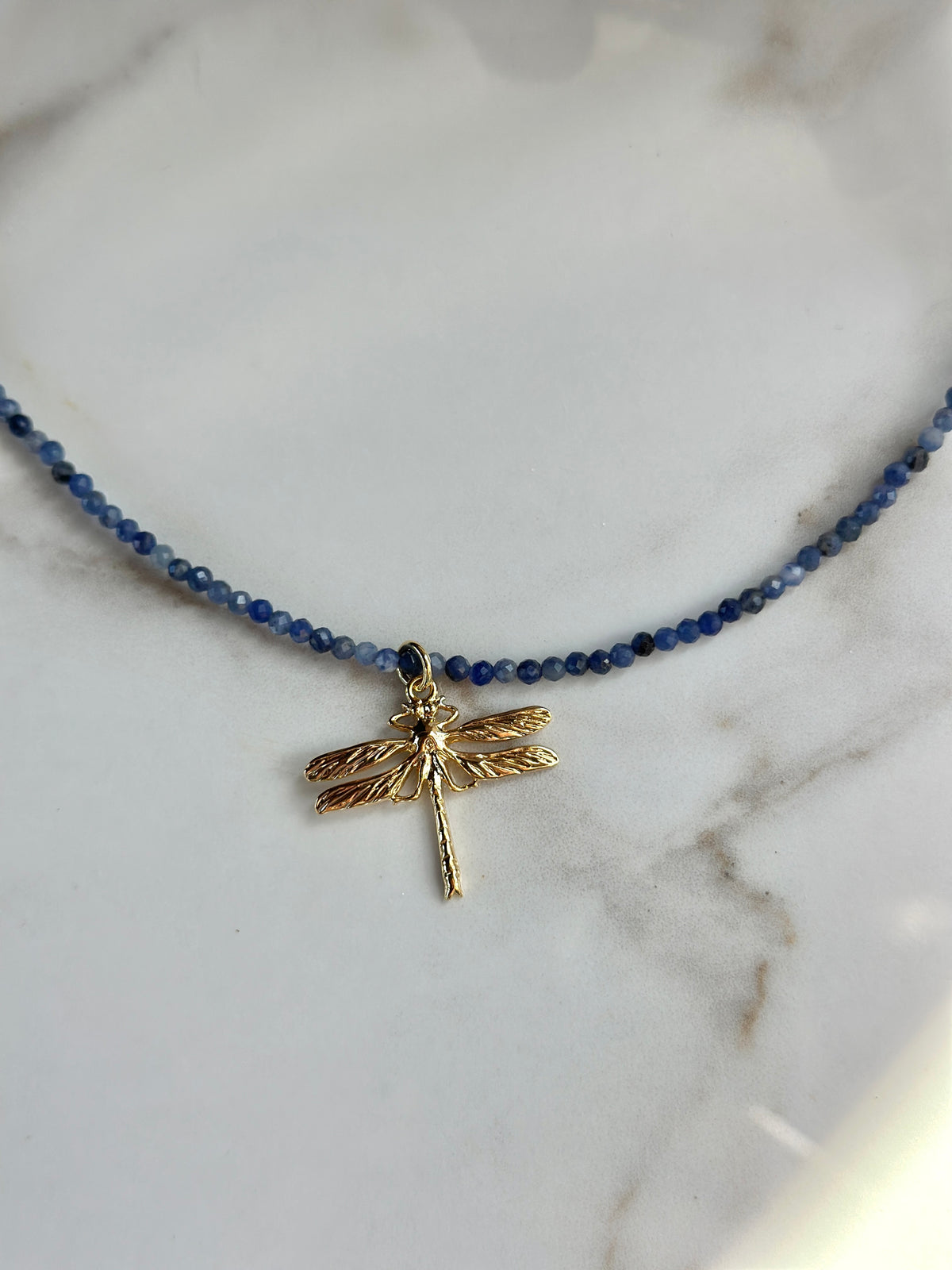 A delicate beaded necklace with small dark blue beads and a gold dragonfly pendant, displayed on a white marble surface.