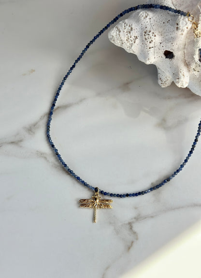 A beaded necklace with deep blue beads and a gold dragonfly pendant, displayed on a white marble surface next to a white seashell.