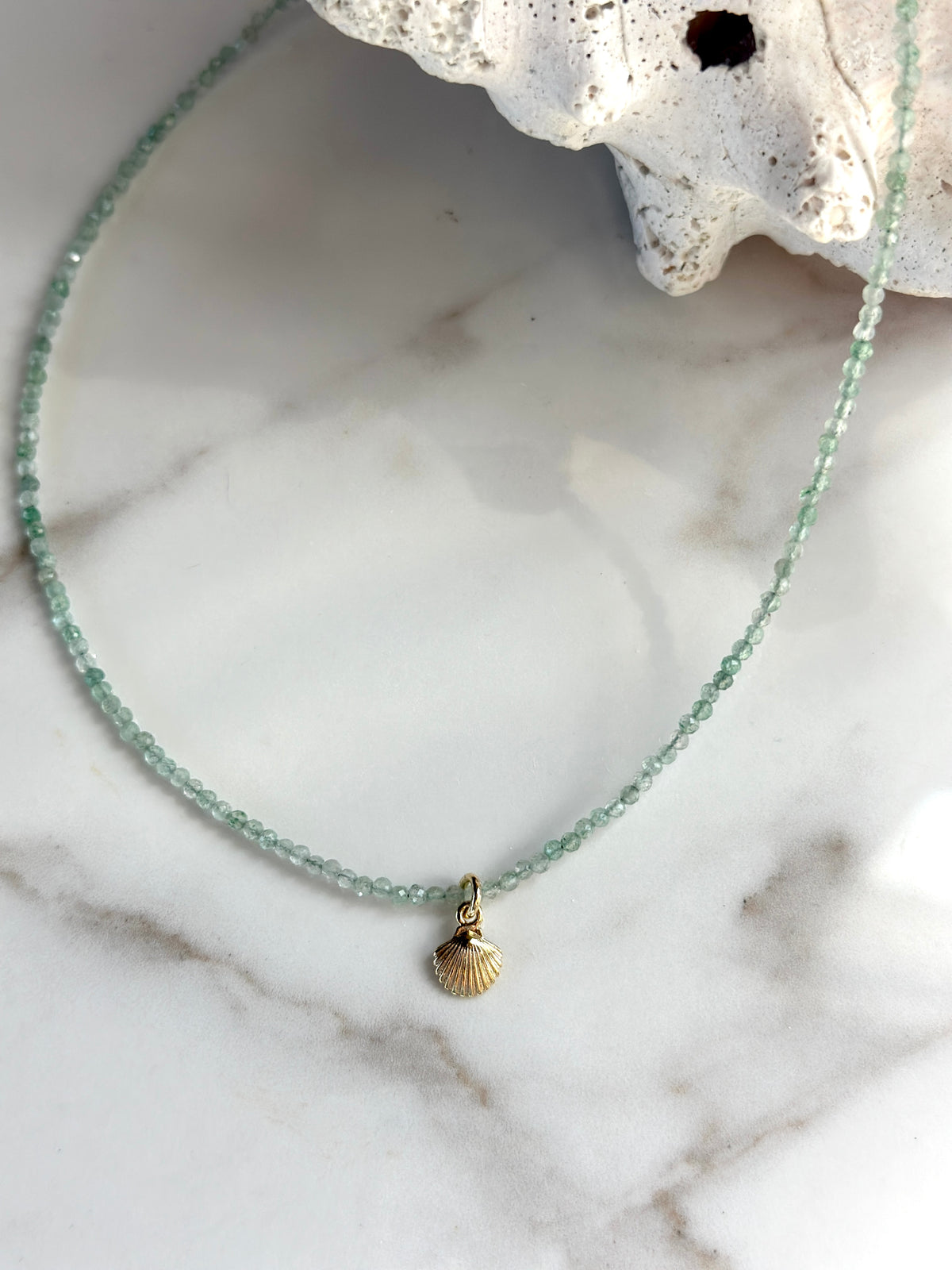 A delicate beaded necklace with light green faceted beads and a small gold seashell pendant, displayed on a white marble surface with a white seashell prop.