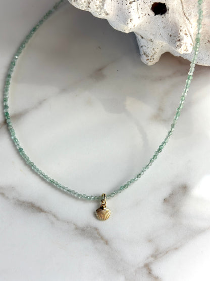 A delicate beaded necklace with light green faceted beads and a small gold seashell pendant, displayed on a white marble surface with a white seashell prop.