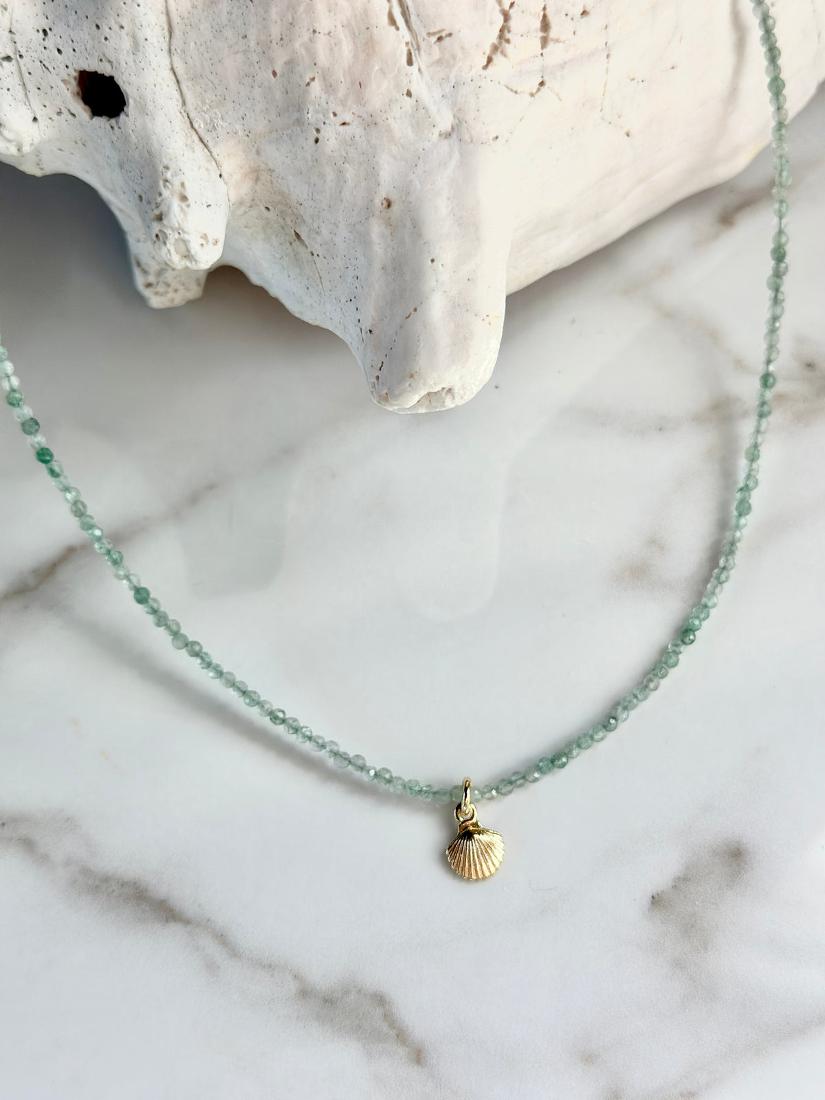 A delicate necklace with a small gold seashell pendant and light green faceted beads, displayed on a white marble surface next to a white seashell prop.