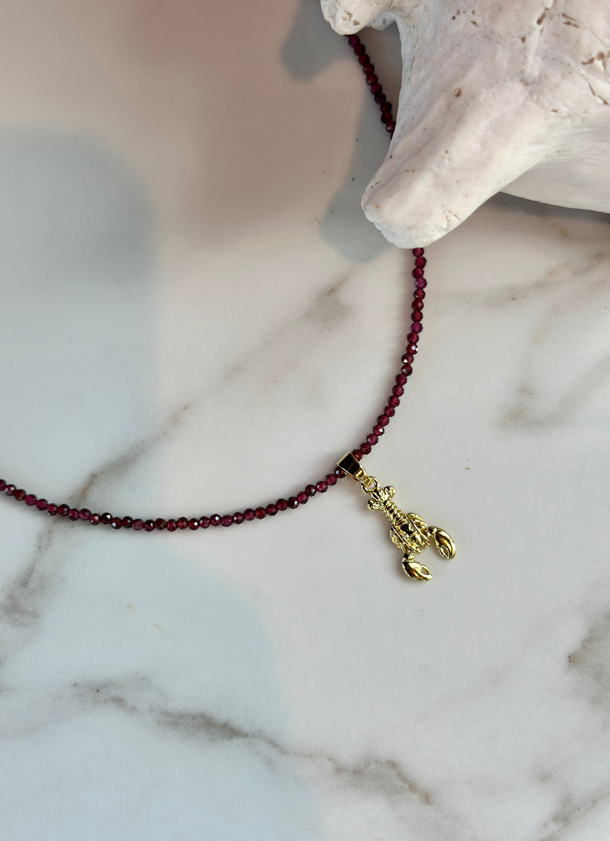 A beaded necklace with dark red faceted beads and a gold lobster charm, displayed on a white marble surface next to a white shell.