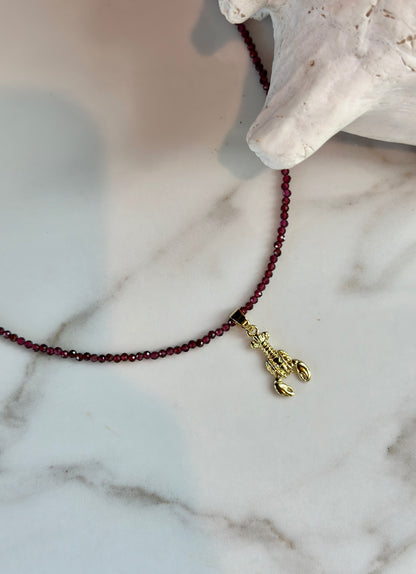 A beaded necklace with dark red faceted beads and a gold lobster charm, displayed on a white marble surface next to a white shell.