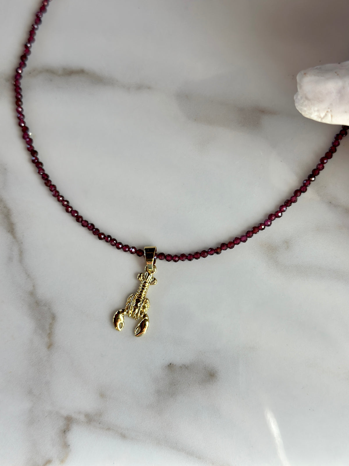 A beaded necklace with dark red garnet faceted beads and a small gold lobster charm, displayed on a white marble surface.