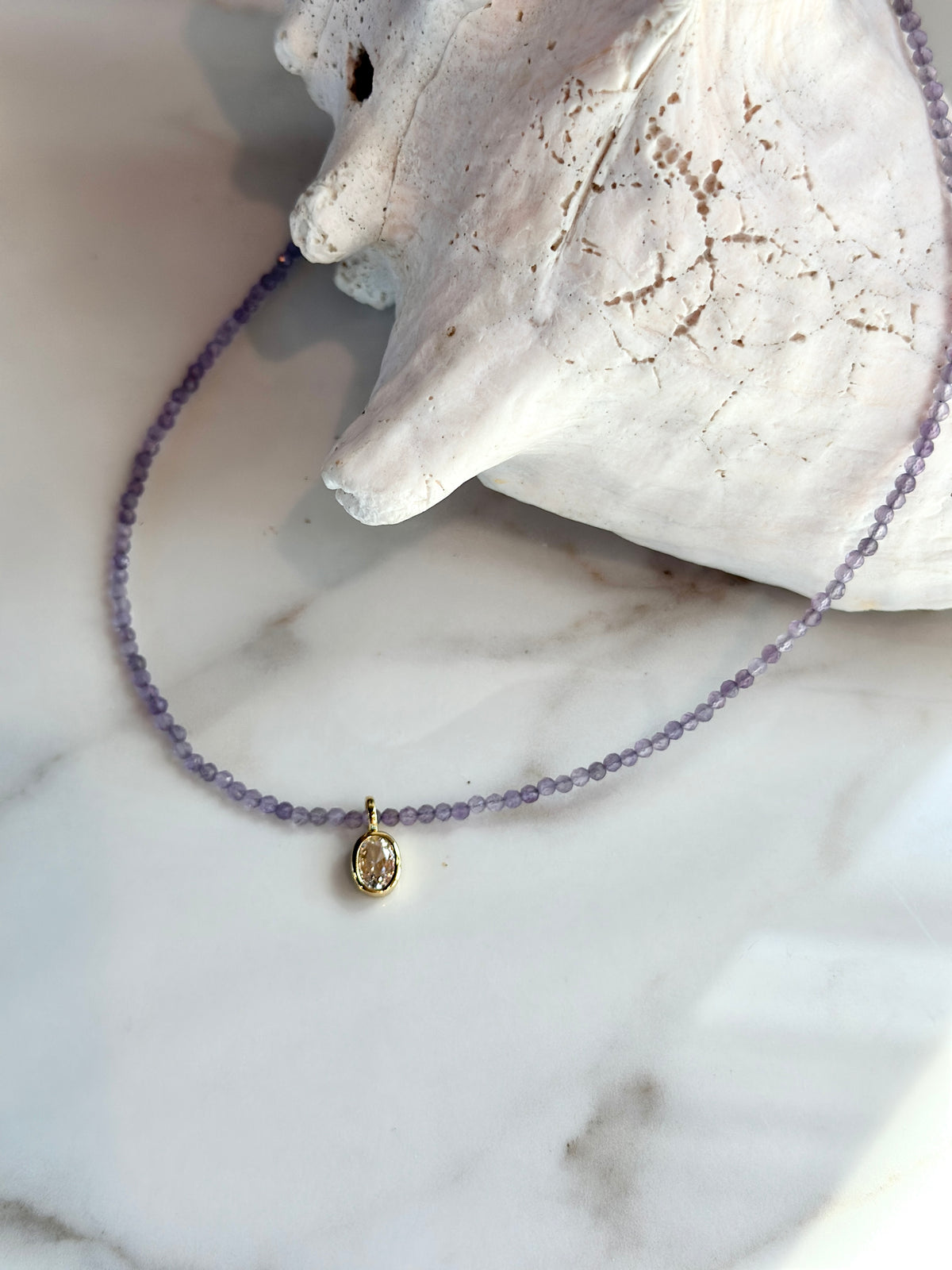 A delicate beaded necklace with small lavender-purple beads and a gold-toned oval pendant, displayed on a white marble surface next to a white seashell.