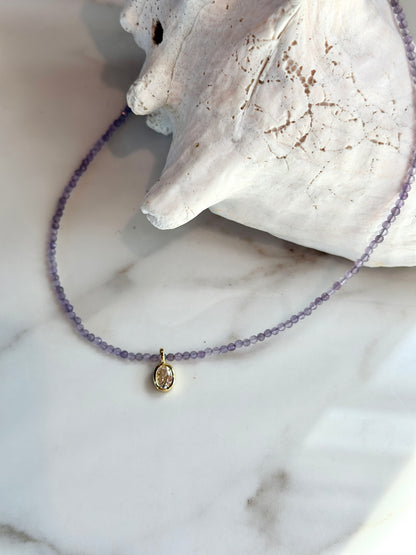 A delicate beaded necklace with small lavender-purple beads and a gold-toned oval pendant, displayed on a white marble surface next to a white seashell.