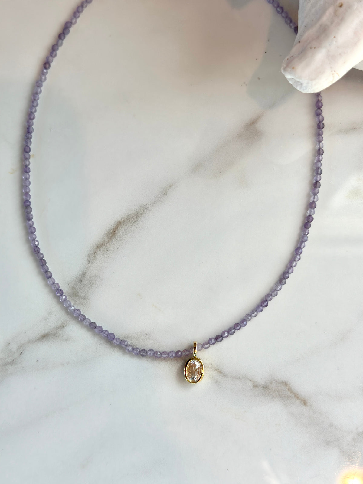 Lavender faceted gemstone bead necklace with a clear oval crystal charm in a gold setting.

