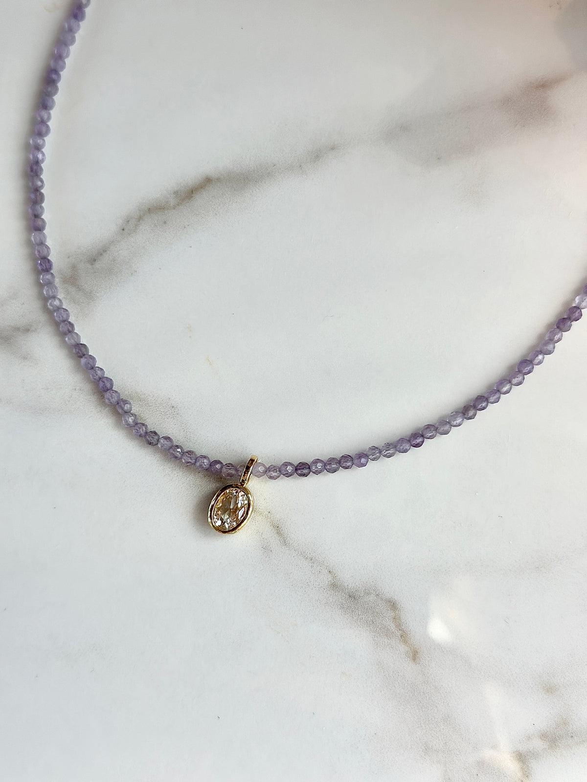 A delicate beaded necklace with small lavender-purple beads and a small oval gold pendant, displayed on a white marble surface.