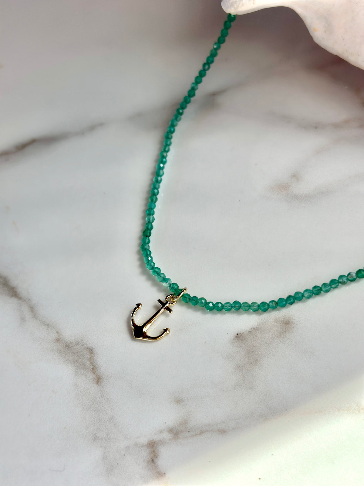 A beaded necklace with small translucent green beads and a gold anchor pendant, displayed on a white marble surface.