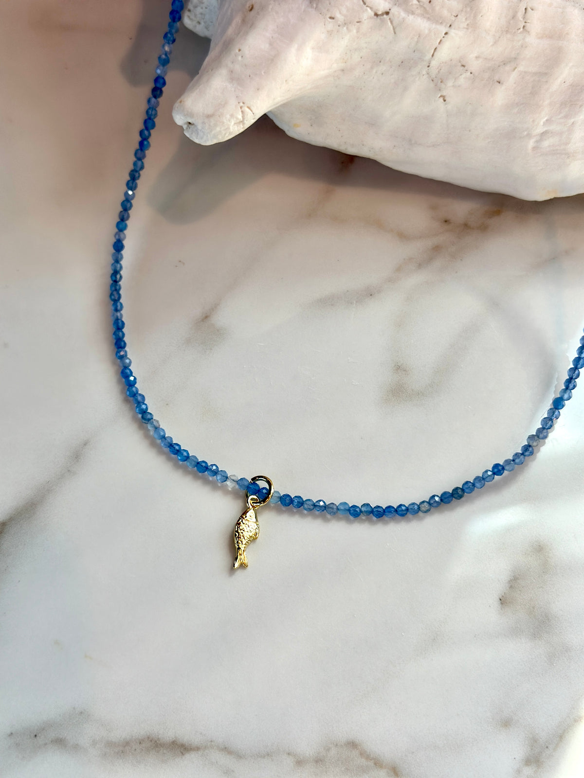A delicate beaded necklace with small blue faceted beads and a small gold seashell pendant, displayed on a white marble surface next to a white seashell.