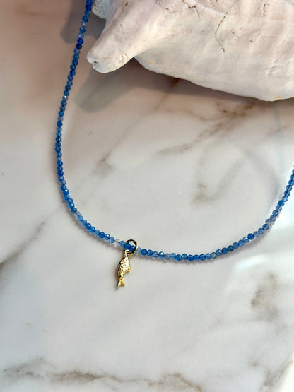 A delicate beaded necklace with small blue faceted beads and a small gold seashell pendant, displayed on a white marble surface next to a white seashell.