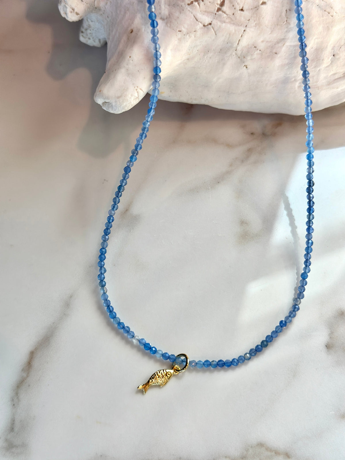 A delicate necklace made of small blue faceted beads with a small gold fish charm, displayed on a white marble surface next to a white seashell.