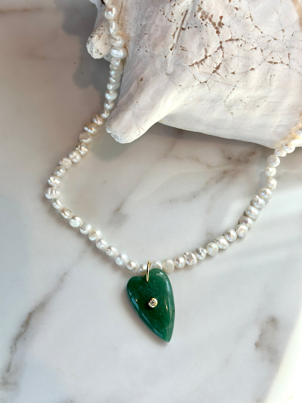 A beaded necklace with white pearl-like beads and a green heart-shaped pendant with a small gold accent, displayed on a white marble surface next to a white shell.