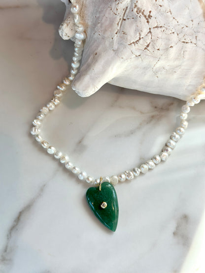 A beaded necklace with white pearl-like beads and a green heart-shaped pendant with a small gold accent, displayed on a white marble surface next to a white shell.